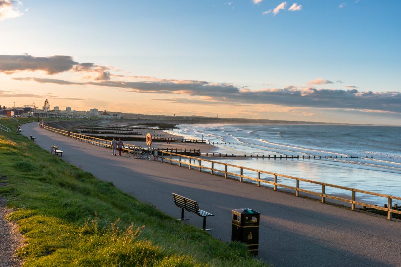 Fenced footpath lined with benches on a seawall along a sandy beach at sunset. People are strolling along the footpath enjoying the magnificent scenery. Aberdeen, Scotland, UK.